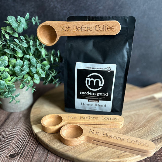 A set of personalized wooden coffee scoops with engraved text 'Not Before Coffee', alongside a Modern Grind coffee bag, on a wooden surface with a small plant decoration in the background.