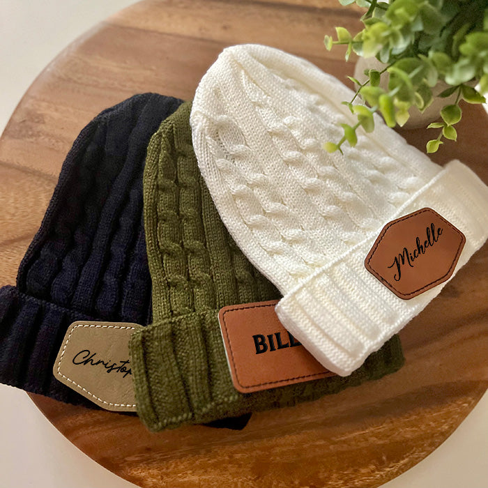 Three knit beanies in black, green, and white colors, each with a customizable leather patch on the fold.