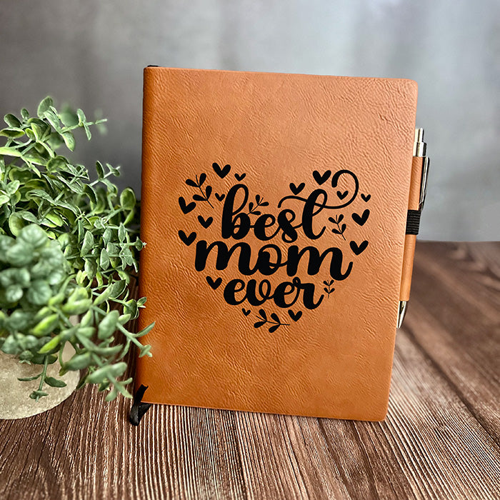 A brown vegan leather notebook with 'best mom ever' engraved on the cover, accompanied by a matching pen, displayed on a wooden surface with a plant decoration.