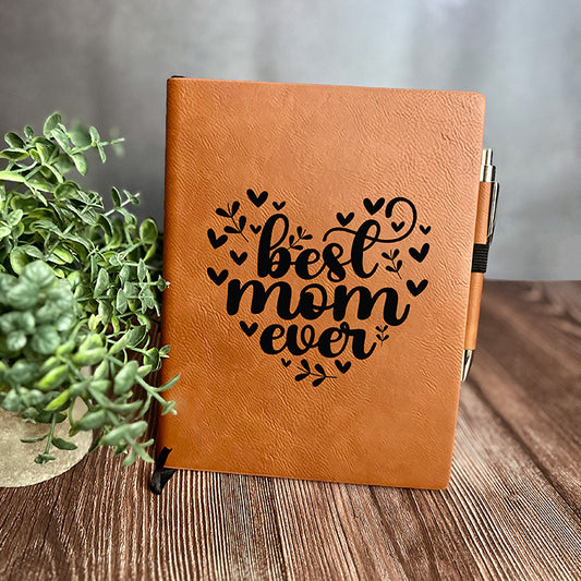A brown vegan leather notebook with 'best mom ever' engraved on the cover, accompanied by a matching pen, displayed on a wooden surface with a plant decoration.