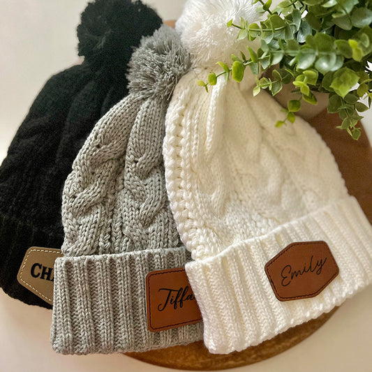 Three cable knit beanies in black, gray, and white colors, each featuring an adorable pom detail and a customizable leather patch with personalized text.