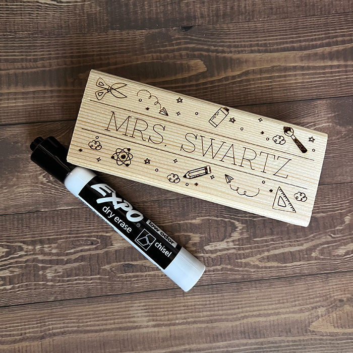 A personalized wooden eraser with engraved teacher's name and a black dry erase marker on a wooden surface.