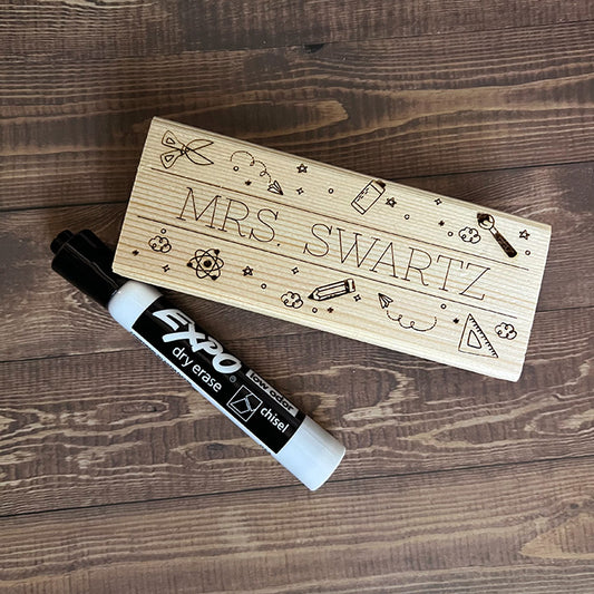 A personalized wooden eraser with engraved teacher's name and a black dry erase marker on a wooden surface.