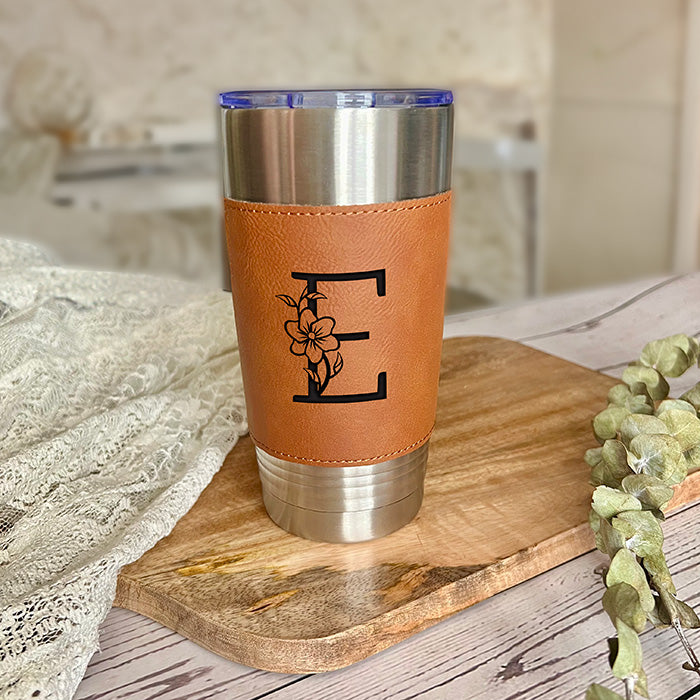 A personalized stainless steel tumbler with a brown leatherette sleeve featuring a custom engraved letter, placed on a wooden board with a decorative flower in the background.
