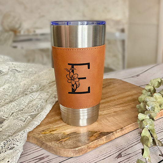 A personalized stainless steel tumbler with a brown leatherette sleeve featuring a custom engraved letter, placed on a wooden board with a decorative flower in the background.