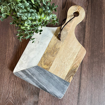 A marble and hard wood cutting board with a personalized laser-etched design, displayed with a wooden handle and a small green plant in the background.