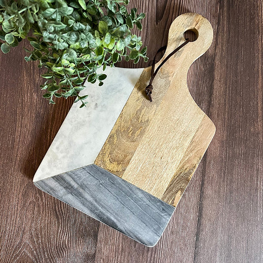 A marble and hard wood cutting board with a personalized laser-etched design, displayed with a wooden handle and a small green plant in the background.