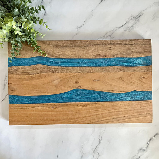 A hard wood cutting board with blue resin accents on the edges, displayed on a marble surface with a small plant in the background.