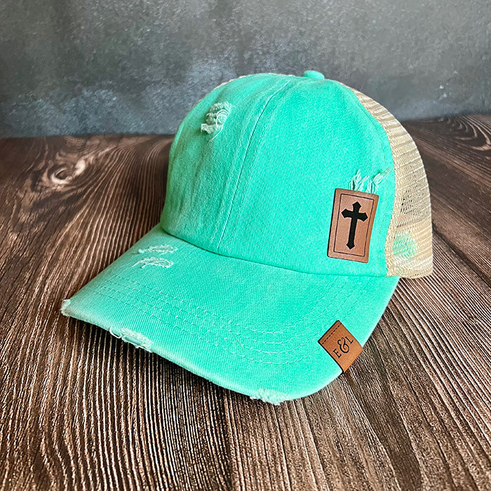 A teal ponytail hat with a Christian cross-etched patch on rawhide vegan leather.