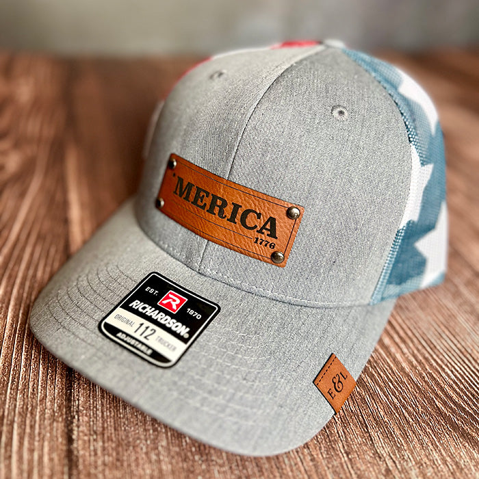 A gray stras and stripes Richardson 112 trucker hat with a leather riveted patch that has '*MERICA' laser-etched on it, displayed on a wooden surface.