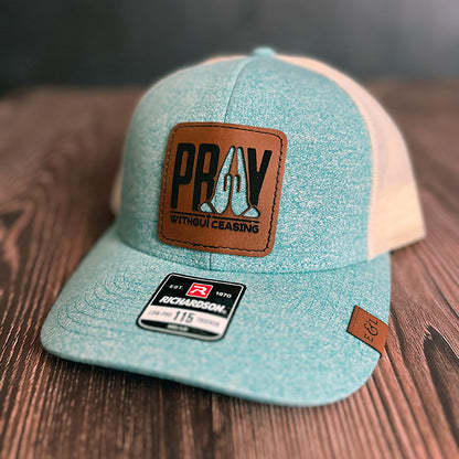 A teal and beige trucker hat with a vegan leather patch that has the text 'PRAY Without Ceasing' and a cutout of praying hands revealing the hat color underneath.